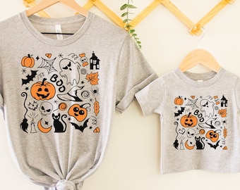 Könnte beinhalten: Zwei graue T-Shirts, eine Erwachsenen- und eine Kindergröße, mit einem Halloween-Motiv. Das Design umfasst orangefarbene Kürbisse, einen Geist, eine schwarze Katze und das Wort "BOO". Die Shirts werden auf Holzbügeln präsentiert.
