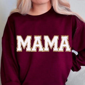May include: Burgundy sweatshirt with the word "MAMA" in white letters with gold glitter outlining the letters.