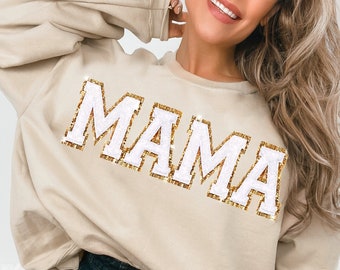 May include: A beige sweatshirt with the word "MAMA" in white letters outlined in gold glitter.