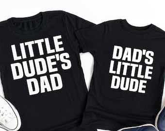 May include: Two black t-shirts with white text. The larger shirt says "Little Dude's Dad" and the smaller shirt says "Dad's Little Dude".