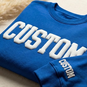 May include: A royal blue sweatshirt with the word "CUSTOM" in large, white, block letters across the chest. The same word is also printed on the sleeve cuff. The sweatshirt is folded on a light-colored surface.