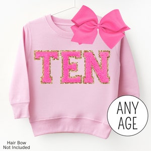 May include: Pink sweatshirt with gold glitter lettering that says "TEN". The sweatshirt is on a hanger and has a pink bow on top.