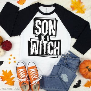 以下が含まれることがあります： 大胆なブロック体のフォントで「SON OF A WITCH」の文字が入った、白と黒のラグランスタイルのシャツ。オレンジ色のスニーカー、ブルージーンズ、カボチャ、葉、クモなどのハロウィーンの装飾が施された画像です。