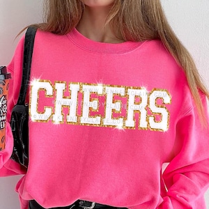 May include: A bright pink sweatshirt with the word "CHEERS" in a gold glitter font. The sweatshirt is a crew neck style. A coffee cup with a coffee bean design is held in the left hand.
