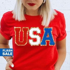 May include: Red, white, and blue glitter "USA" graphic on a red t-shirt. The shirt is being held up by a person wearing a red, white, and blue striped hat.