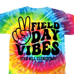 Puede incluir: Una camiseta te&ntilde;ida con un dise&ntilde;o de nudo con un signo de la paz y las palabras "FIELD DAY VIBES" en negro. La camiseta tambi&eacute;n incluye el texto "STAR HILL ELEMENTARY" en negro. La camiseta es una mezcla de colores del arco&iacute;ris.
