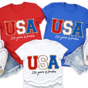 May include: Three t-shirts in red, white, and blue with the word "USA" in sequined letters. Each shirt has the text "250 years of freedom" below the word "USA". The shirts are paired with denim shorts, white sneakers, and small American flags.