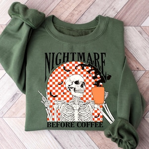 May include: Green sweatshirt with the text "NIGHTMARE BEFORE COFFEE" above a skeleton graphic holding a coffee cup. The graphic includes bats and a checkered orange and white background. The sweatshirt is folded on a wooden surface.