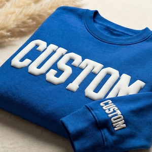 May include: A royal blue sweatshirt with the word "CUSTOM" in white, block letters across the chest. The same word is also printed on the sleeve cuff. The sweatshirt is folded on a white surface.
