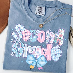 May include: Blue jean colored t-shirt with the words "Second Grade" in a colorful, patterned design. The design includes bows, flowers, and a pencil. A gold necklace with a pendant is draped across the shirt.
