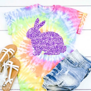 May include: A tie-dye t-shirt with a purple glitter bunny silhouette. The shirt is paired with denim shorts and white sandals.