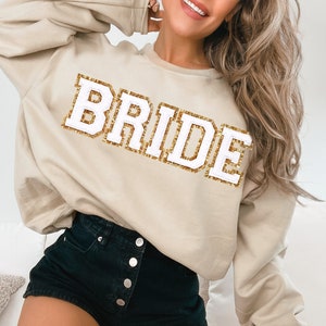 May include: A beige sweatshirt with the word "BRIDE" in white letters outlined in gold glitter.