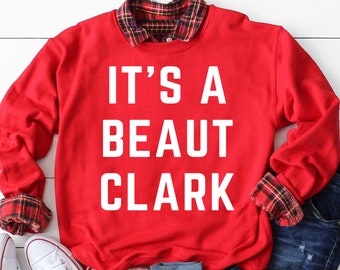以下が含まれることがあります： 白い文字で「IT'S A BEAUT CLARK」と書かれた赤いスウェットシャツ。スウェットシャツは、チェック柄のシャツの上に重ねて、青いジーンズと合わせています。