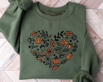 Könnte beinhalten: Grünes Sweatshirt mit einem herzförmigen Design, das mit herbstlichen Illustrationen wie Kürbissen, Blättern und Blumen gefüllt ist. Der Text "GREEN SWEATSHIRT" befindet sich unten im Bild. Das Sweatshirt liegt auf einer Holzoberfläche.