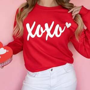 May include: A red sweatshirt with the white text "XOXO" and a white heart. A person is holding a red heart-shaped box with the word "LOVE" and an arrow through a heart. The background is a solid pink color.