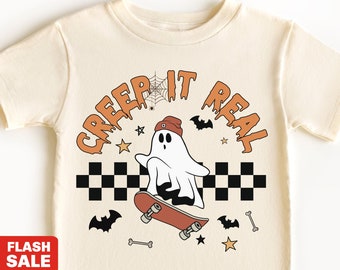 May include: Cream-colored natural tee shirt with a Halloween-themed graphic. The design features a ghost on a skateboard, the phrase "CREEP IT REAL", bats, stars, and a checkered pattern. The shirt is a natural tee.