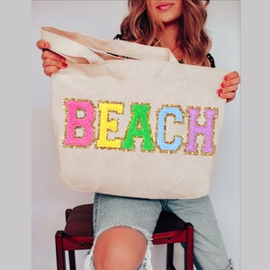 May include: A white canvas tote bag with the word "BEACH" spelled out in colorful, glittery letters. The letters are pink, yellow, green, light blue, and purple.