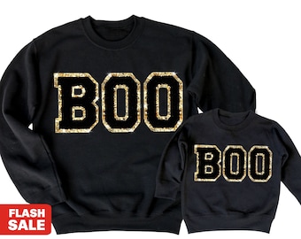 May include: Black sweatshirts featuring the word "BOO" in gold glitter. The matching set includes an adult and a child's size, ideal for Halloween or themed events. The glitter adds a festive touch.