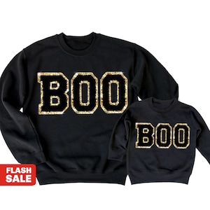 May include: Black sweatshirts with the word "BOO" in gold glitter lettering. The sweatshirts are a matching set, one adult size and one child size, perfect for Halloween or a spooky themed event.
