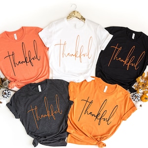 May include: Five t-shirts in various colors: orange, white, black, dark gray, and a lighter orange. Each shirt has the word "Thankful" in a cursive font. The shirts are displayed on a white surface with fall-themed decorations.