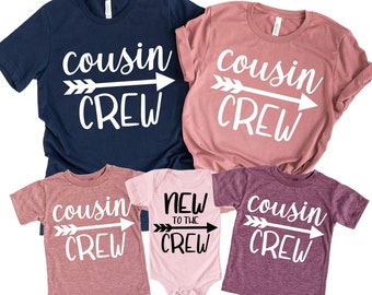 May include: Set of four t-shirts with the text "cousin crew" and an arrow graphic. The shirts are in navy blue, pink, and purple. One shirt says "new to the crew".