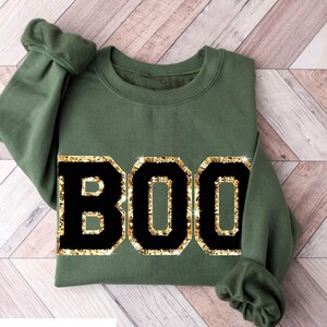 May include: Green sweatshirt with the word "BOO" in large black letters outlined in gold glitter. The text is centered on the front of the sweatshirt. The bottom of the image reads "GREEN SWEATSHIRT".