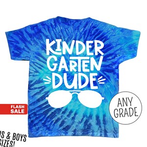 Op de afbeelding: Blauw tie-dye T-shirt met de witte woorden "KINDERGARTEN DUDE" en een witte zonnebrilafbeelding. Het shirt heeft ook de woorden "ANY GRADE" in een witte cirkel. Een rode banner leest "FLASH SALE".
