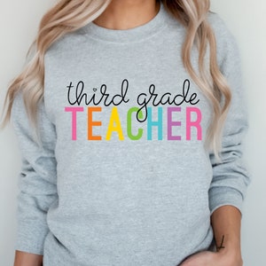 Puede incluir: Una sudadera gris con el texto "third grade TEACHER" en colores arco&iacute;ris.