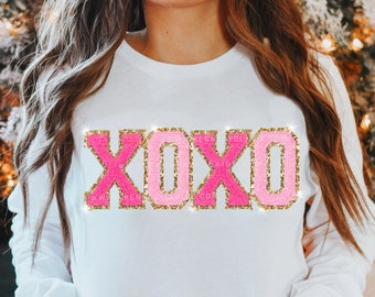 May include: White long-sleeve shirt with the letters XOXO in pink and gold glitter patches. The shirt has a crew neck and is made with real patches. The letters are outlined with gold glitter.