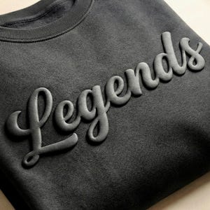 May include: A charcoal gray crewneck sweatshirt featuring the word Legends in a raised, three-dimensional, puff-style script font across the chest. The garment has a ribbed collar and a soft, textured fabric finish with a consistent dark tone.