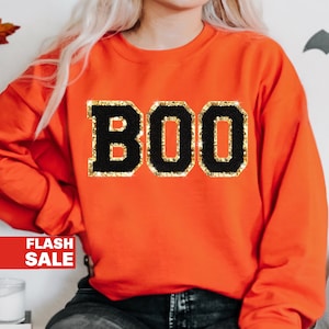 May include: An orange sweatshirt with the word "BOO" in large, black, glitter-edged letters. The sweatshirt is a crew neck style, perfect for Halloween or fall. The image also includes a "FLASH SALE" banner.