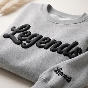 May include: A gray sweatshirt with the word "Legends" in large, raised black lettering across the chest. The sleeve cuff also features the word "Legends" in black. The sweatshirt is made of a soft material.