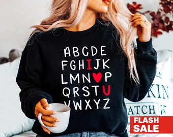 Może przedstawiać: Czarna bluza z białymi literami alfabetu, które piszą "I U" z czerwonym sercem. Bluza ma okrągły dekolt. Obraz zawiera również biały kubek i słowa "FLASH SALE" w czerwonym banerze.