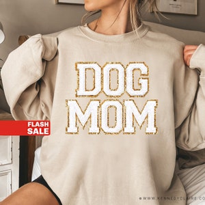 May include: Beige sweatshirt with the text "DOG MOM" in white block letters outlined with gold glitter. The sweatshirt has long sleeves and a crew neck. A red banner with the words "FLASH SALE" is in the lower left corner.