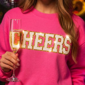 May include: A bright pink sweatshirt with the word "CHEERS" in white letters outlined with gold glitter. A person holds a champagne flute filled with bubbly liquid. The background has bokeh lights.