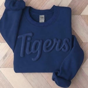 May include: A navy blue crewneck sweatshirt featuring the word Tigers in a raised, tonal puff print across the chest. The garment has long sleeves with ribbed cuffs and a Gildan brand label visible at the inner neckline, showcasing a solid color.