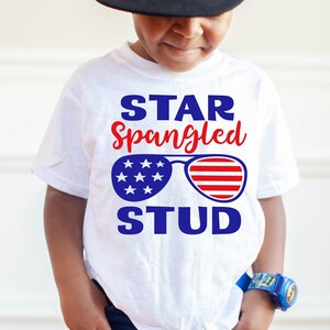 May include: White t-shirt with a blue and red design. The design features the text "STAR Spangled STUD" with a pair of sunglasses with an American flag pattern.