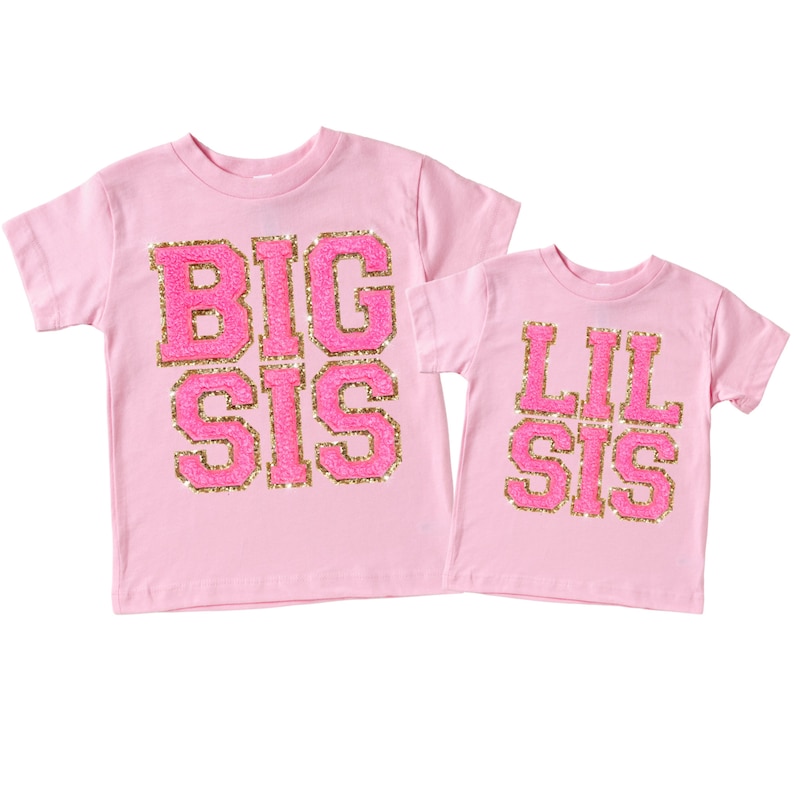 May include: Two pink t-shirts with gold glitter lettering. The larger shirt says "BIG SIS" and the smaller shirt says "LIL SIS".