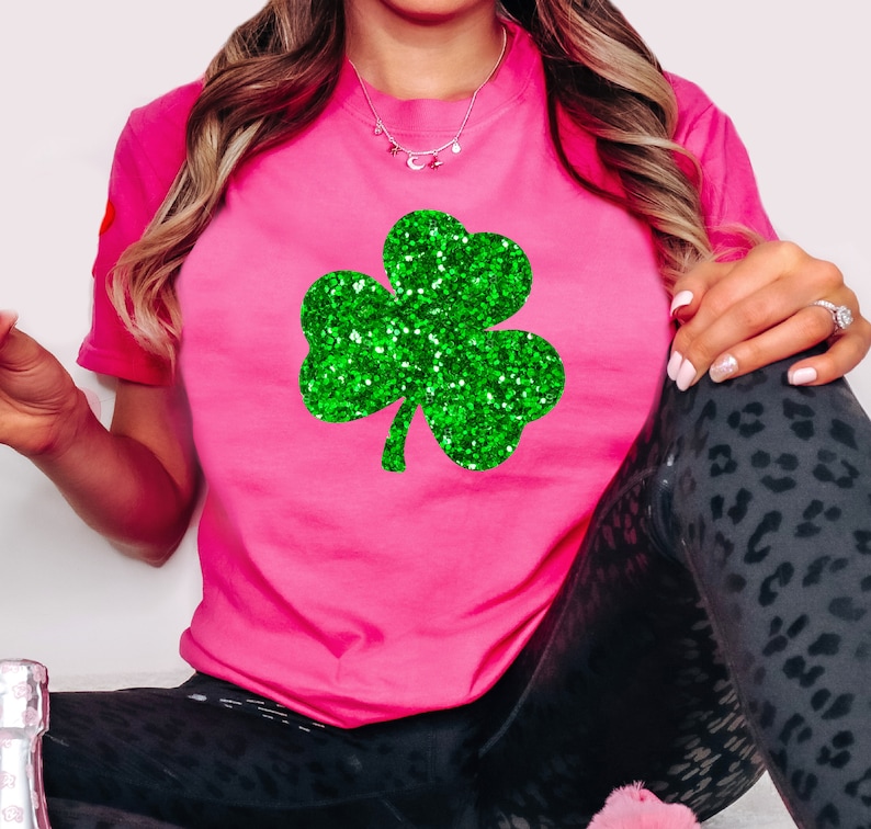 May include: A bright pink t-shirt featuring a large, glittery green shamrock design. The shamrock is centered on the shirt, creating a festive and eye-catching look. The shirt is short-sleeved and made of a soft material.