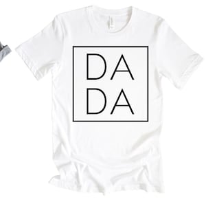 Puede incluir: Camiseta blanca con un cuadrado negro que contiene el texto "DA DA" en una fuente en negrita sin serif.