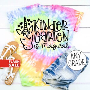 Op de afbeelding: Een tie-dye T-shirt met een eenhoorn en de tekst "Kindergarten is magical". Het shirt is gecombineerd met een spijkerbroekje en sandalen. De tekst "Any Grade" is zichtbaar in een cirkel rechtsonder in de afbeelding.