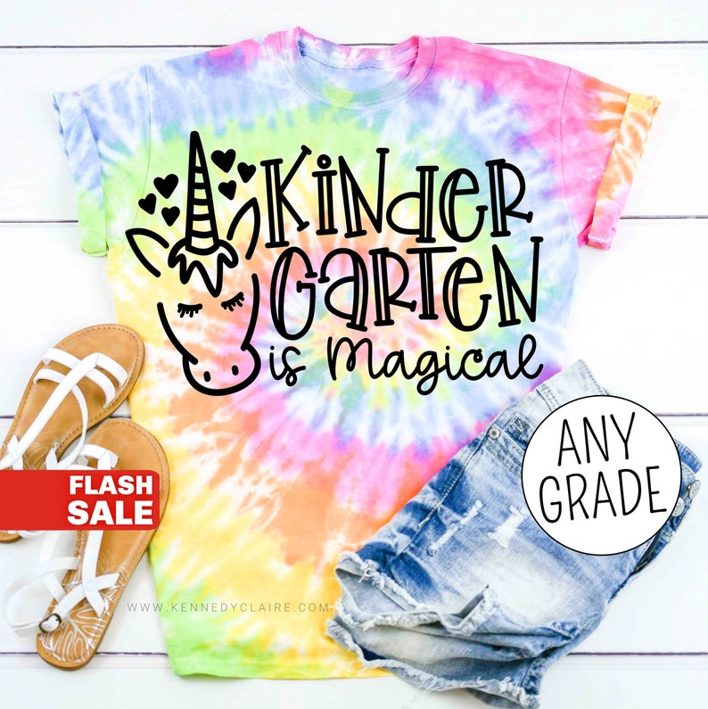 Op de afbeelding: Een tie-dye T-shirt met een eenhoorn en de tekst "Kindergarten is magical". Het shirt is gecombineerd met een spijkerbroekje en sandalen. De tekst "Any Grade" is zichtbaar in een cirkel rechtsonder in de afbeelding.