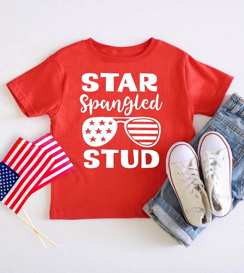 May include: Red short-sleeved t-shirt with a white graphic that says "Star Spangled Stud" with a pair of sunglasses with stars and stripes. The sunglasses are white with red and blue accents.