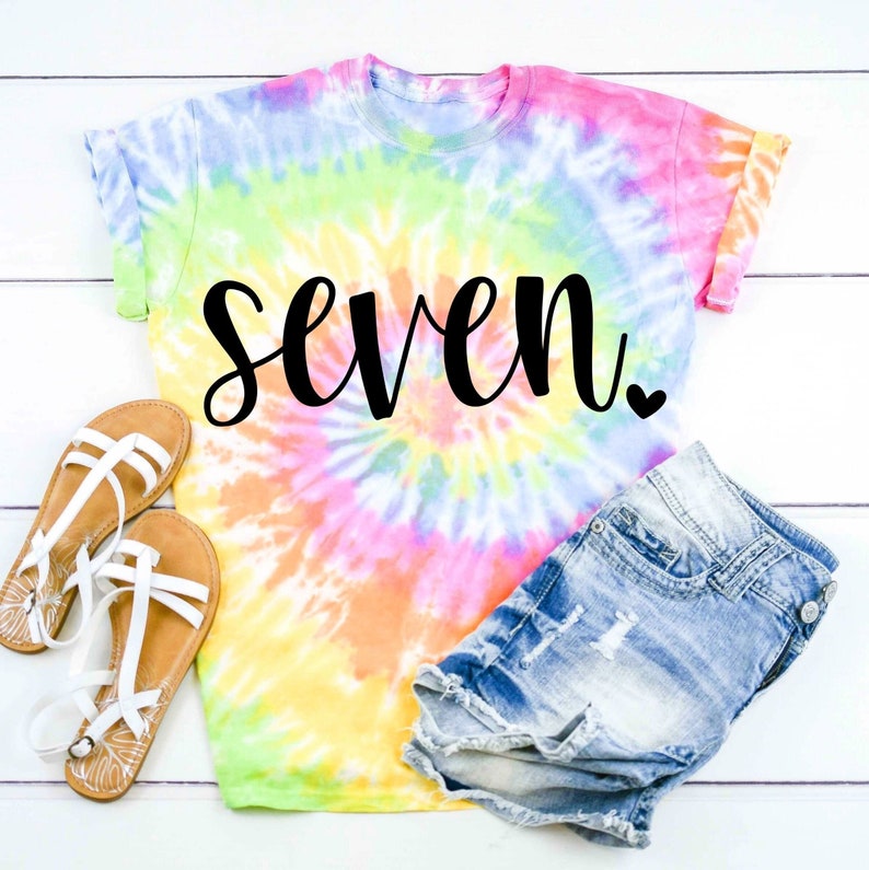 May include: A tie-dye t-shirt with the word "seven" in black script and a small heart. The shirt is a light pink, yellow, and blue color.