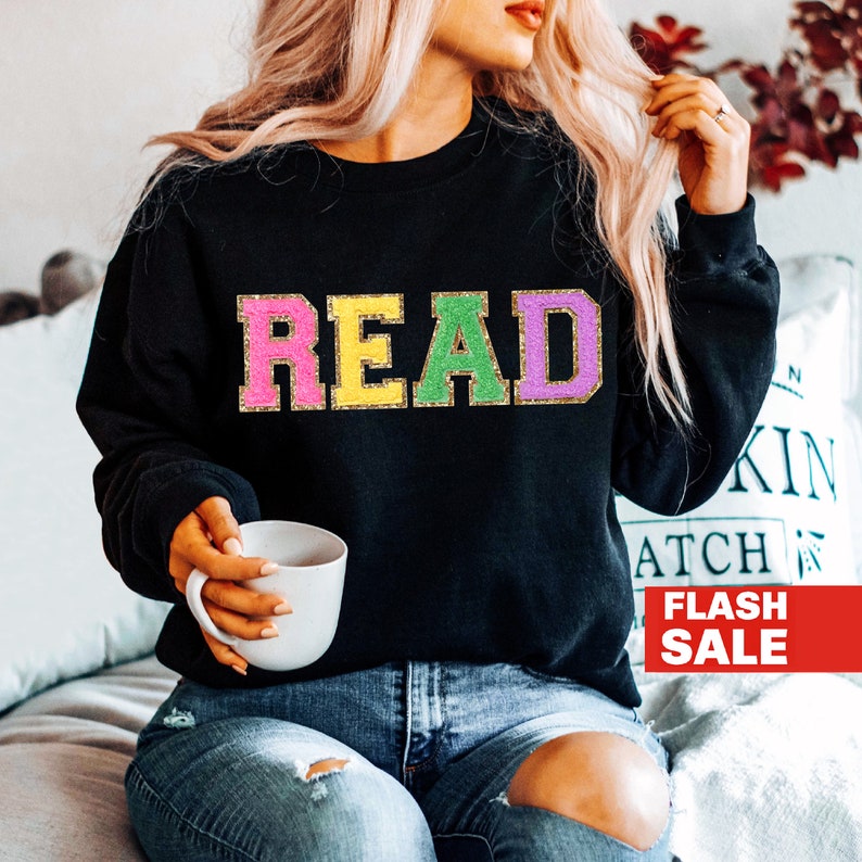 May include: A black sweatshirt with the word "READ" in colorful letters with gold glitter outlining. The letters are pink, yellow, green, and purple. The sweatshirt is being worn by a person sitting on a couch.