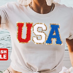 May include: White t-shirt with the word "USA" in red, white, and blue, with gold glitter trim. The letters are in a varsity style. A red "FLASH SALE" banner is in the lower left corner.