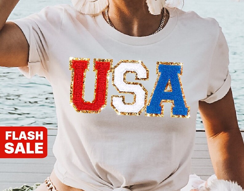 May include: White t-shirt with the word "USA" in red, white, and blue, with gold glitter trim. The letters are in a varsity style. A red "FLASH SALE" banner is in the lower left corner.