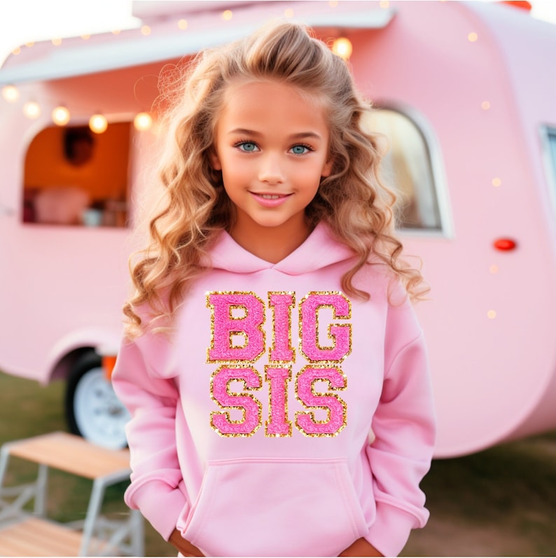 May include: A young girl wearing a pink hoodie with the words "BIG SIS" in gold glitter lettering. The hoodie has a drawstring hood and a kangaroo pocket.