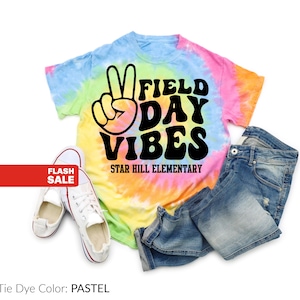Puede incluir: Una camiseta te&ntilde;ida con tinte pastel con las palabras "FIELD DAY VIBES" y un signo de la paz. El texto "STAR HILL ELEMENTARY" est&aacute; debajo. La imagen tambi&eacute;n incluye un par de zapatillas blancas y unos pantalones cortos vaqueros.