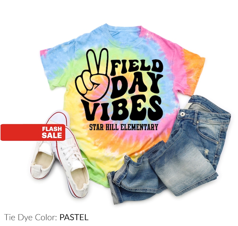 Puede incluir: Una camiseta te&ntilde;ida con tinte pastel con las palabras "FIELD DAY VIBES" y un signo de la paz. El texto "STAR HILL ELEMENTARY" est&aacute; debajo. La imagen tambi&eacute;n incluye un par de zapatillas blancas y unos pantalones cortos vaqueros.